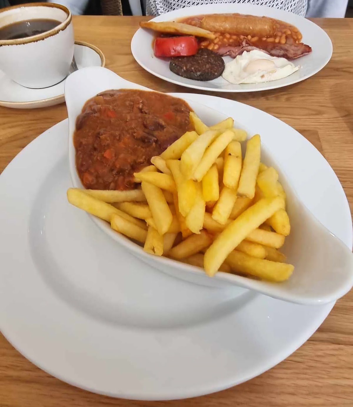 A hearty stew-like bowl with chips on a white plate, with a breakfast plate and a black coffee behind.